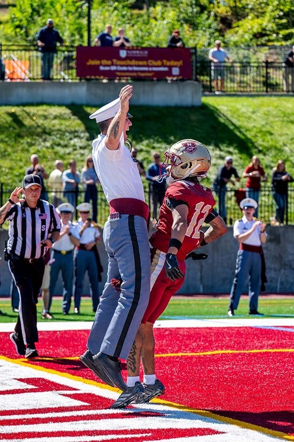 Norwich football player #10 jumps with a uniformed cadet in the end zone.