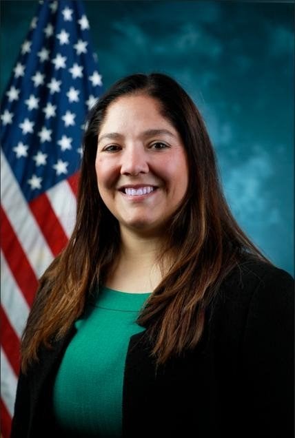 A person in a black blazer poses in front of a U.S. flag and a blue-green backdrop.