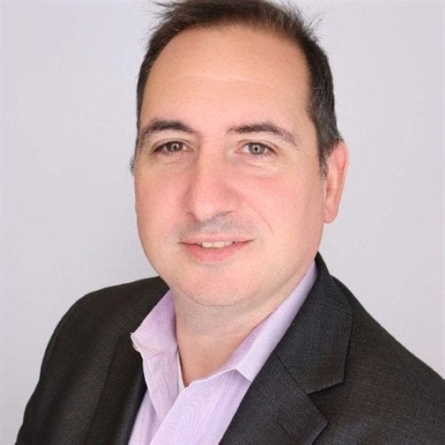 A person in a dark blazer and light collared shirt poses against a light gray background.