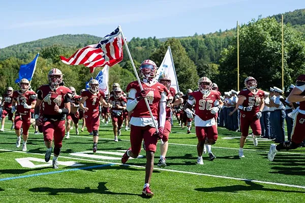 Norwich football players run onto the field as player #4 carries an American flag.