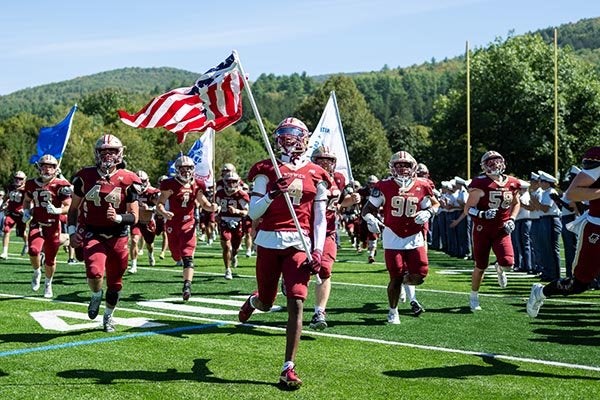 Norwich football players run onto the field as player #4 carries an American flag.