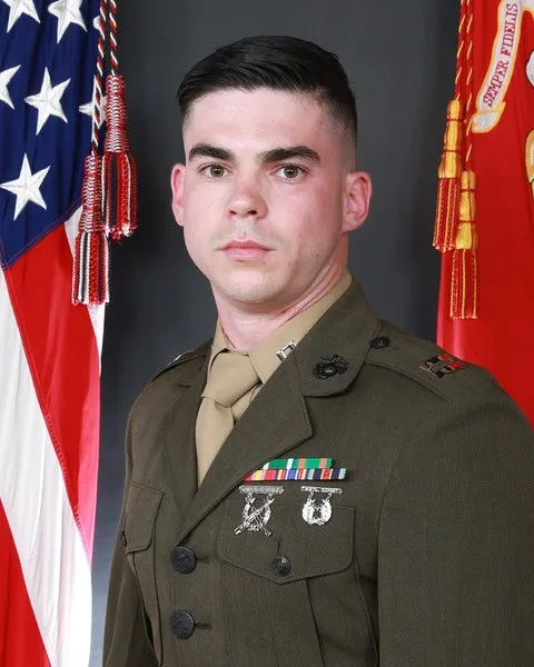 A service member in dress uniform poses between a U.S. flag and a red military flag.