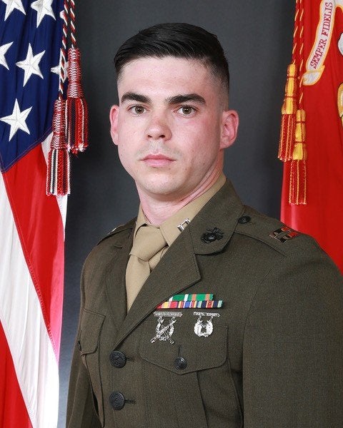 A service member in dress uniform poses between a U.S. flag and a red military flag.