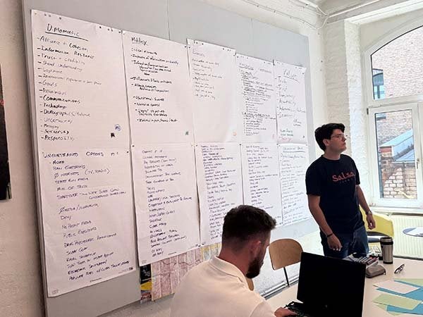 Two people work in a room with large sheets of handwritten notes covering the wall.