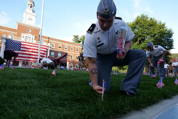 Cadets place small U.S. flags in the grass in front of a large U.S. flag and campus building.