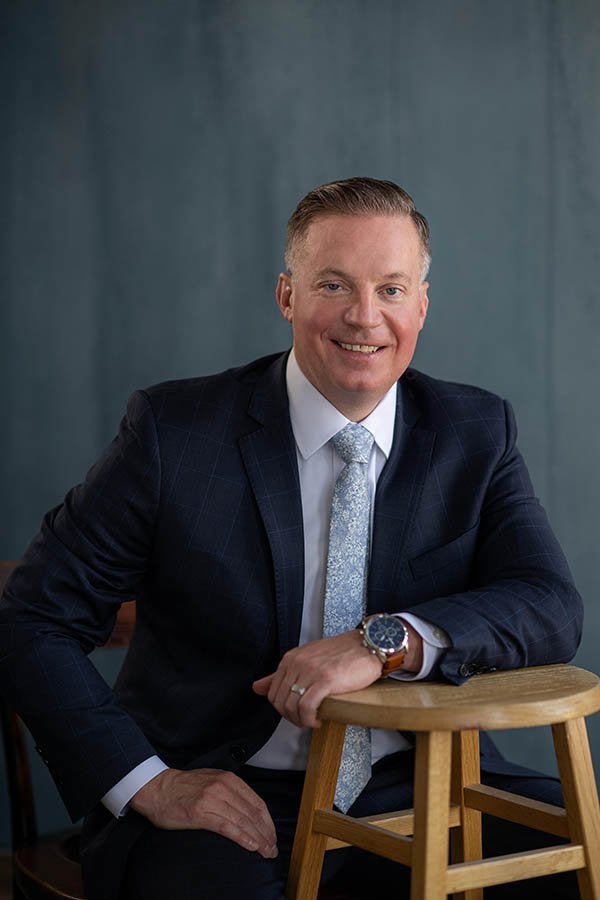 A person in a dark suit and patterned tie sits beside a wooden stool against a blue-gray background.