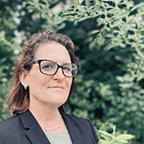 Person wearing glasses and a dark blazer stands in front of leafy trees.