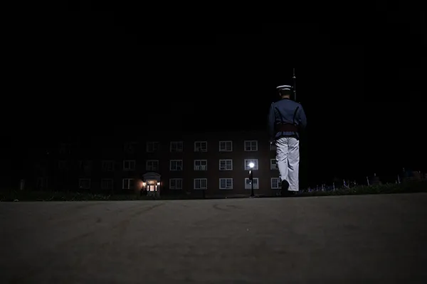 Cadet stands at attention on a dark walkway outside a campus building at night.
