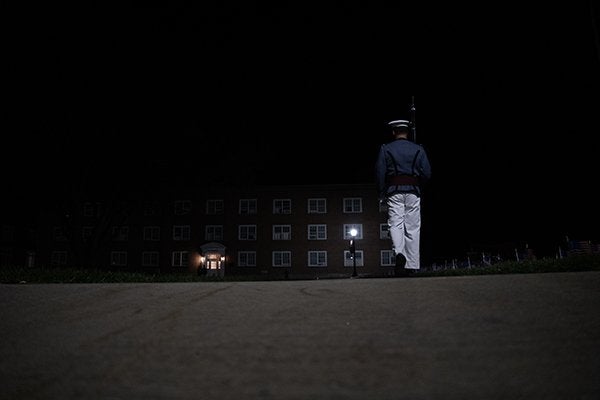 Cadet stands at attention on a dark walkway outside a campus building at night.