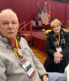 Two people wearing event badges and medals sit in front of a stage with an American flag and Norwich banners.