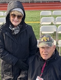 Two people wearing dark jackets pose together in front of folding chairs beside an outdoor athletic field.