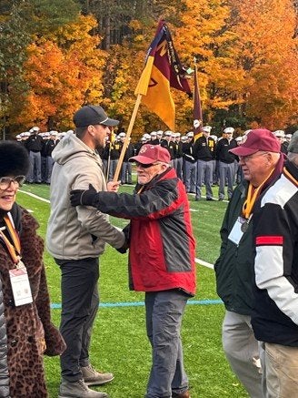 A person holding a Norwich flag greets another person on an athletic field as uniformed cadets stand in formation behind them.