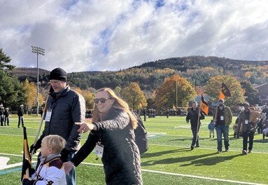 A person wearing sunglasses gestures across an athletic field as a child and other attendees walk nearby carrying flags.