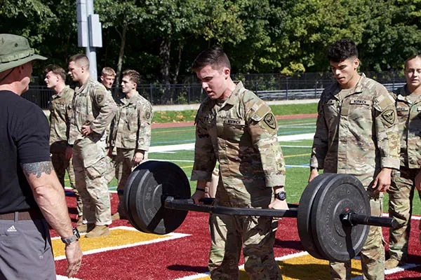 Cadet lifts a loaded barbell on an athletic field as other cadets wait nearby.