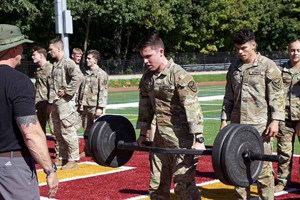 Cadet lifts a loaded barbell on an athletic field as other cadets wait nearby.