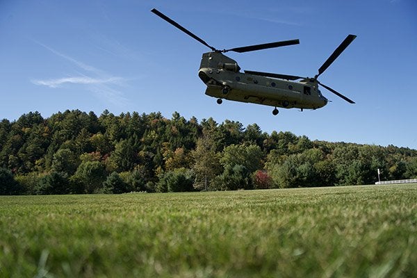 A military Chinook helicopter flies low over a grassy field with a wooded hillside in the background.