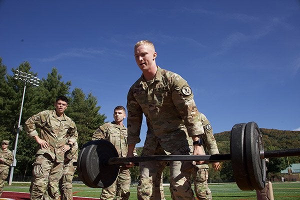 A Texas A&M cadet in camouflage uniform lifts a weighted barbell on a football field as other cadets watch.