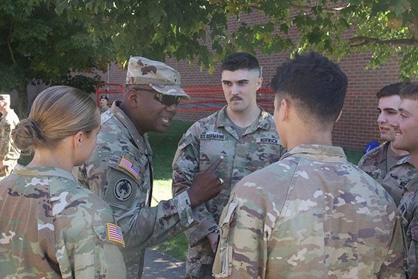 A service member in camouflage uniform gestures while speaking with a group of Norwich cadets outdoors.