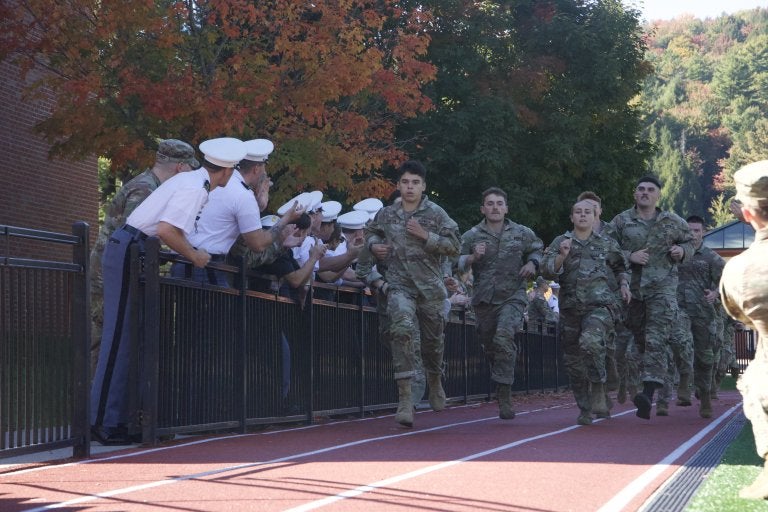 Norwich cadets in camouflage uniforms run along a track as cadets in white shirts and caps cheer from behind a railing.