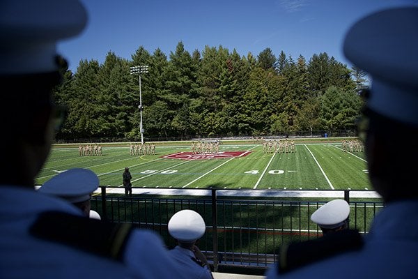 Uniformed cadets in white caps watch groups of cadets standing in formation on a football field.