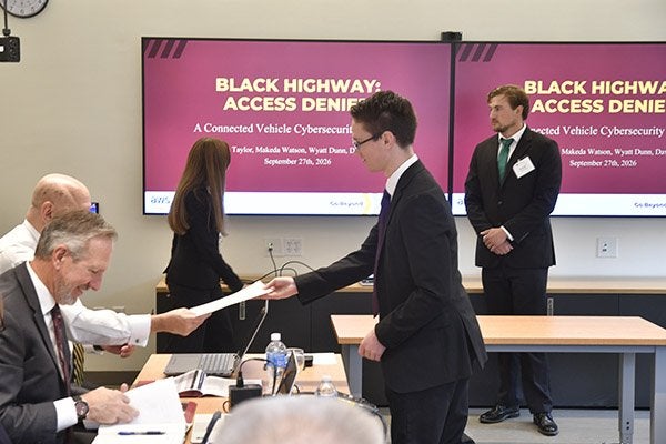 Presenter hands a paper to a seated attendee as screens display a connected vehicle cybersecurity presentation.
