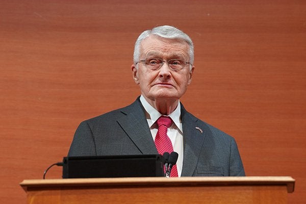 A person with gray hair and glasses stands behind a wooden podium with microphones, wearing a dark suit and red tie.
