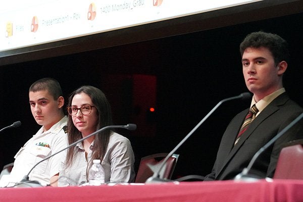Three panelists sit behind microphones at a table, including a service member in a white uniform.
