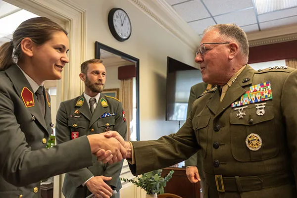 Two service members in military dress uniforms shake hands indoors as another service member stands nearby.
