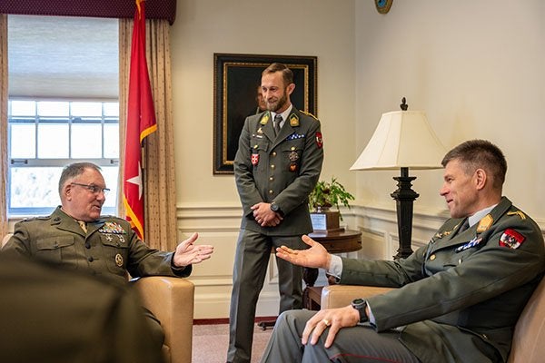 Two service members in dress uniforms sit facing each other and gesture during a conversation as a third stands nearby.