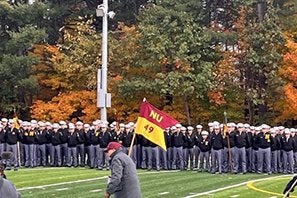 A person walks across a football field carrying a red and gold NU flag as uniformed cadets stand in formation behind them.