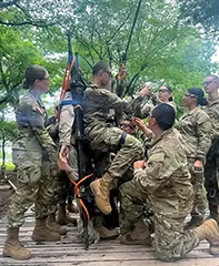 Service members in camouflage uniforms help a person suspended in a harness adjust ropes during an outdoor training exercise.