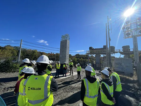 A group wearing hard hats and reflective safety vests gathers at a construction site with concrete pillars and scaffolding.