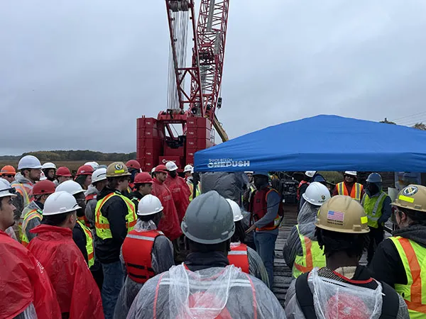 A group wearing hard hats and safety gear gathers around a speaker beneath a blue tent at a construction site.