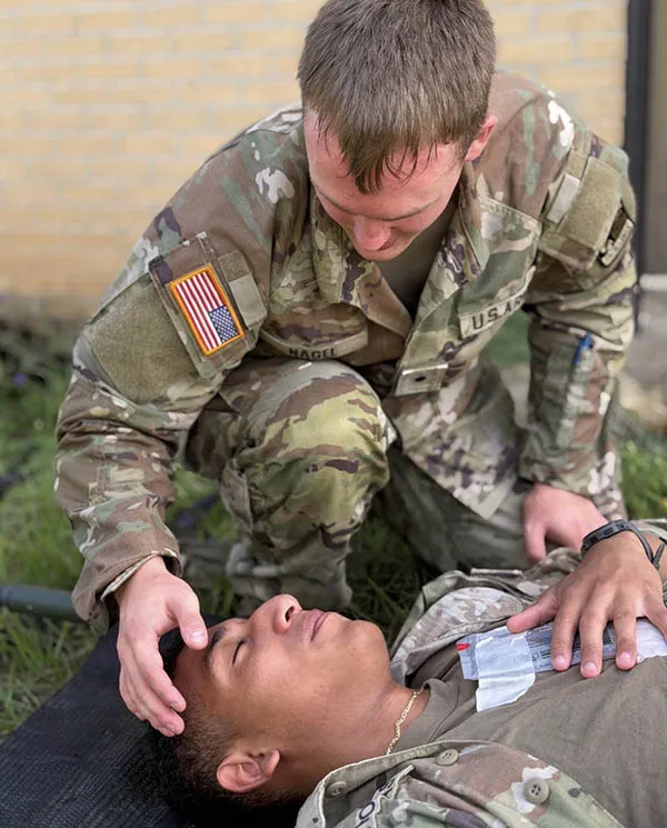 Service member in camouflage uniform kneels beside another service member lying on a mat and supports their head.