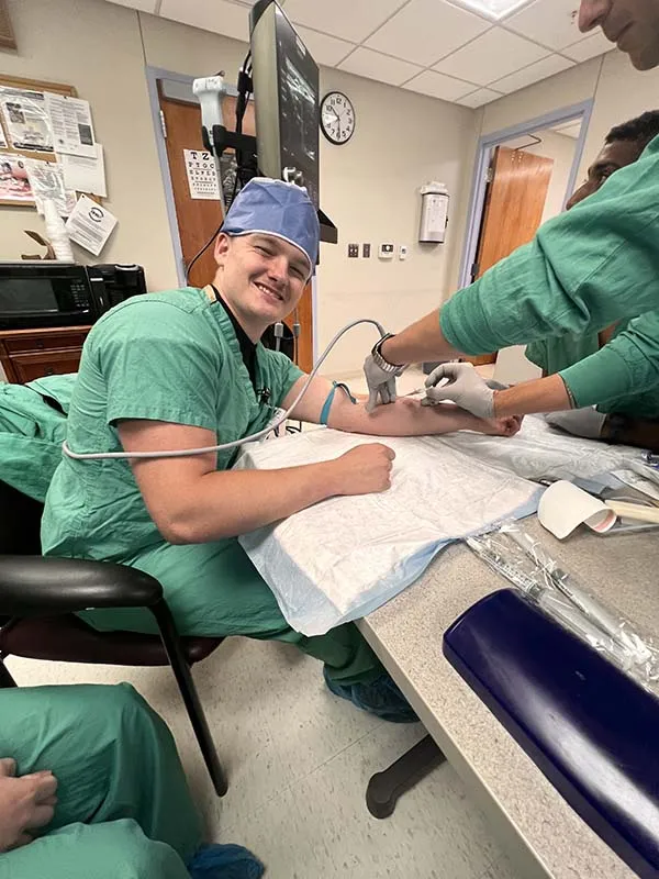 A healthcare worker in green scrubs uses an ultrasound probe and needle on another healthcare worker's arm.