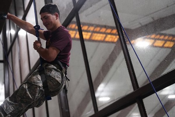 Cadet wearing a safety harness climbs an indoor obstacle while holding a rope.