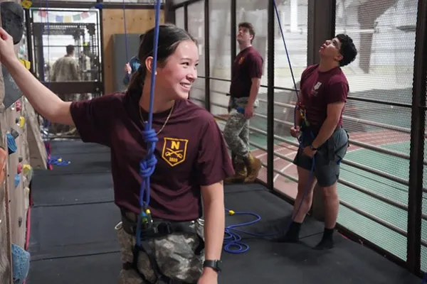 Cadets wearing safety harnesses stand beside an indoor climbing wall with ropes.