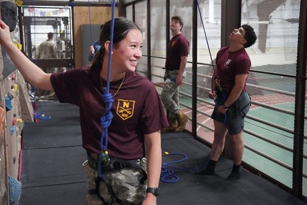 Cadets wearing safety harnesses stand beside an indoor climbing wall with ropes.