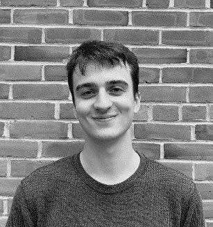 A person with short dark hair wearing a dark sweater poses in front of a brick wall.