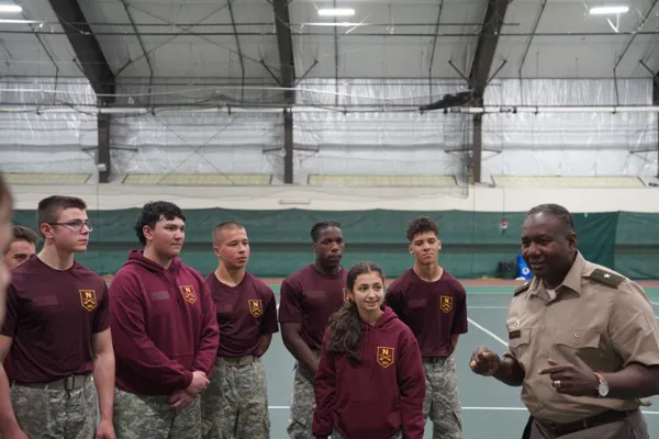 Service member speaks to a group of cadets gathered inside an athletic facility.