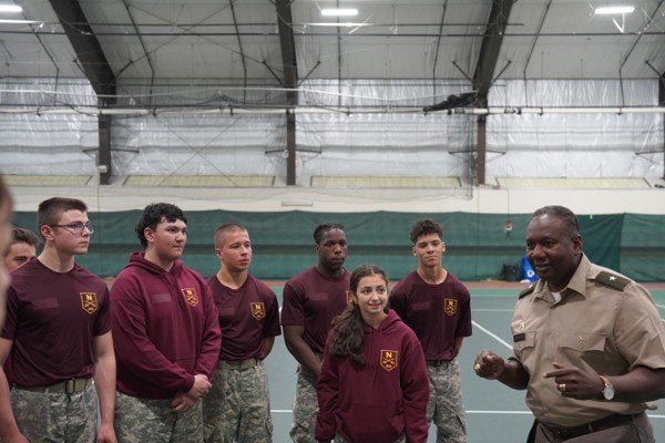 Service member speaks to a group of cadets gathered inside an athletic facility.