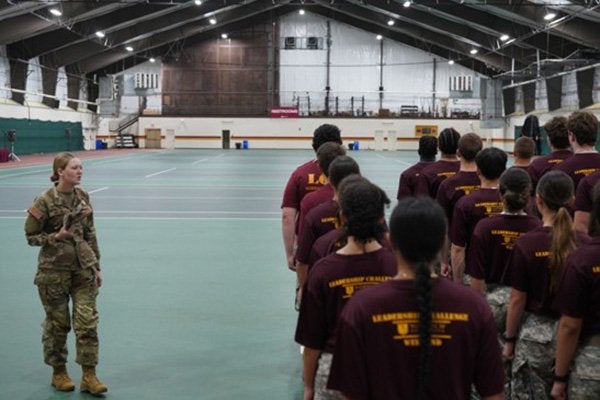 Service member speaks to cadets standing in formation inside an athletic facility.