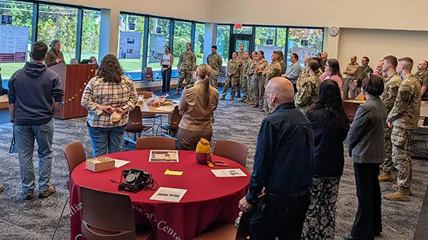 People stand around tables during a presentation as service members line the back of the room.