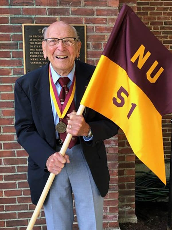 Portrait of R. Damon holding a maroon and gold Norwich University Class of 1951 flag while wearing a medal.