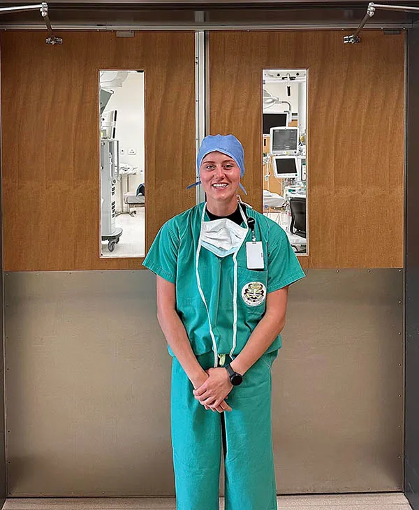 A healthcare worker in green surgical scrubs and a blue cap stands in front of operating room doors.