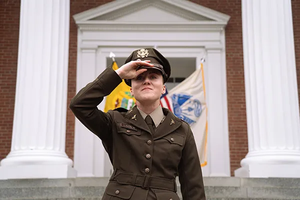 Service member in brown Army dress uniform adjusts their cap in front of a brick building with white columns and flags.