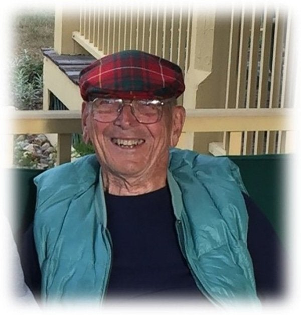 A person wearing glasses, a red plaid cap, and a teal vest smiles while seated outdoors.