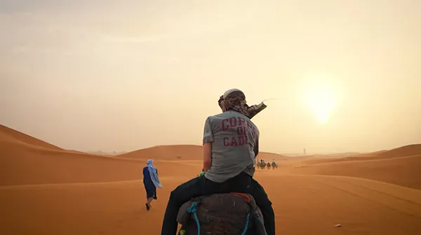 A person wearing a Corps of Cadets shirt rides a camel across desert dunes toward a group of riders at sunset.