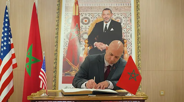 A person in a suit signs a guest book at a desk with U.S. and Moroccan flags beneath a large framed portrait.