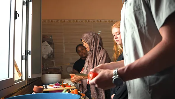 A group prepares vegetables in a kitchen, with one person cutting a tomato and another slicing vegetables near a window.